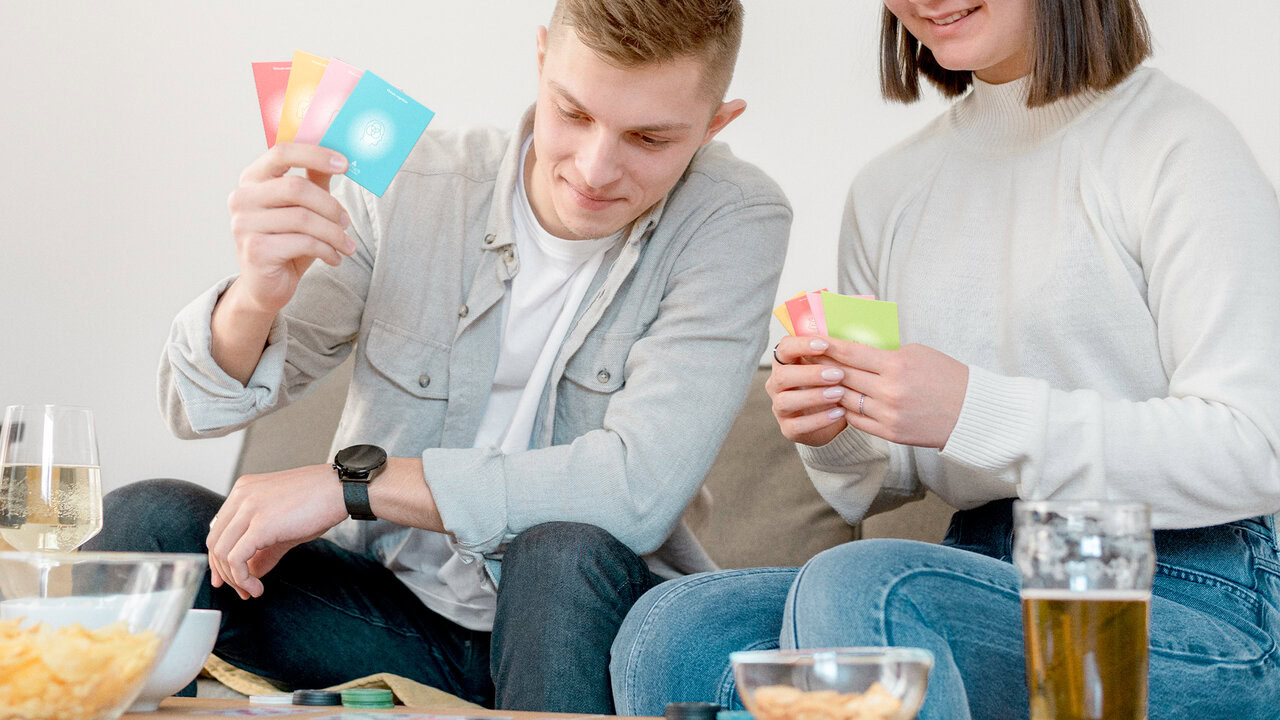 Nebly_Encartados_pareja_con_las_cartas_en_lamano Hombre y mujer sosteniendo cartas de 'Encartados', un juego de cartas diseñado para actividad en pareja. Sentados en un ambiente informal con bebidas y aperitivos.