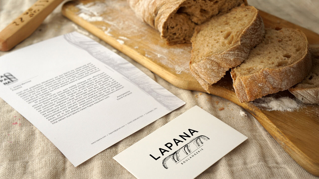 Nebly_LAPANA_TarjetaHoja_1920x1080 Elementos de papelería corporativa de LAPANA (tarjeta de presentación, hoja membretada) junto a un pan artesanal. Diseño de identidad para panadería artesanal.