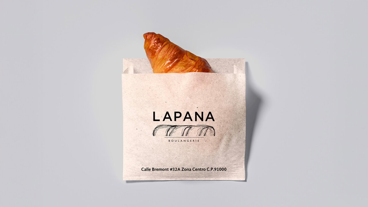 Nebly_LAPANA_bolsa_1920x1080 Bolsa de papel de con un croissant asomando. El diseño de empaque incluye el logotipo de LAPANA y su dirección. Branding acogedor para panadería artesanal: identidad cálida y tradición europea.