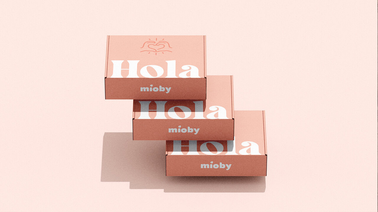 Nebly_Mioby_cajas Cajas de envío de Mioby, apiladas. El diseño de empaque es en color rosa claro con el texto Hola y un logotipo de manos formando un corazón.
