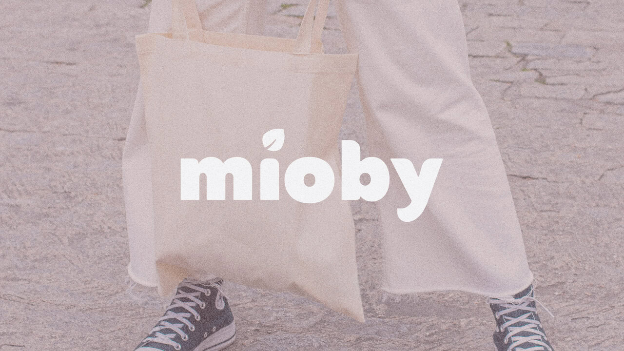 Nebly_Mioby_logo Persona de pie, vistiendo pantalones blancos y zapatillas, sosteniendo una tote bag con el logotipo de la marca impreso en la bolsa. Identidad única para emprendedores: piezas hechas a mano y esencia cálida.