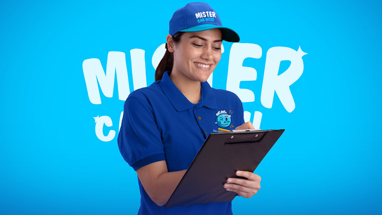 Nebly-Mister-Carwash-uniforme Mujer joven sonriente con una playera y gorra azul con el logo de mister car wash mientras escribe algo en un tablero.