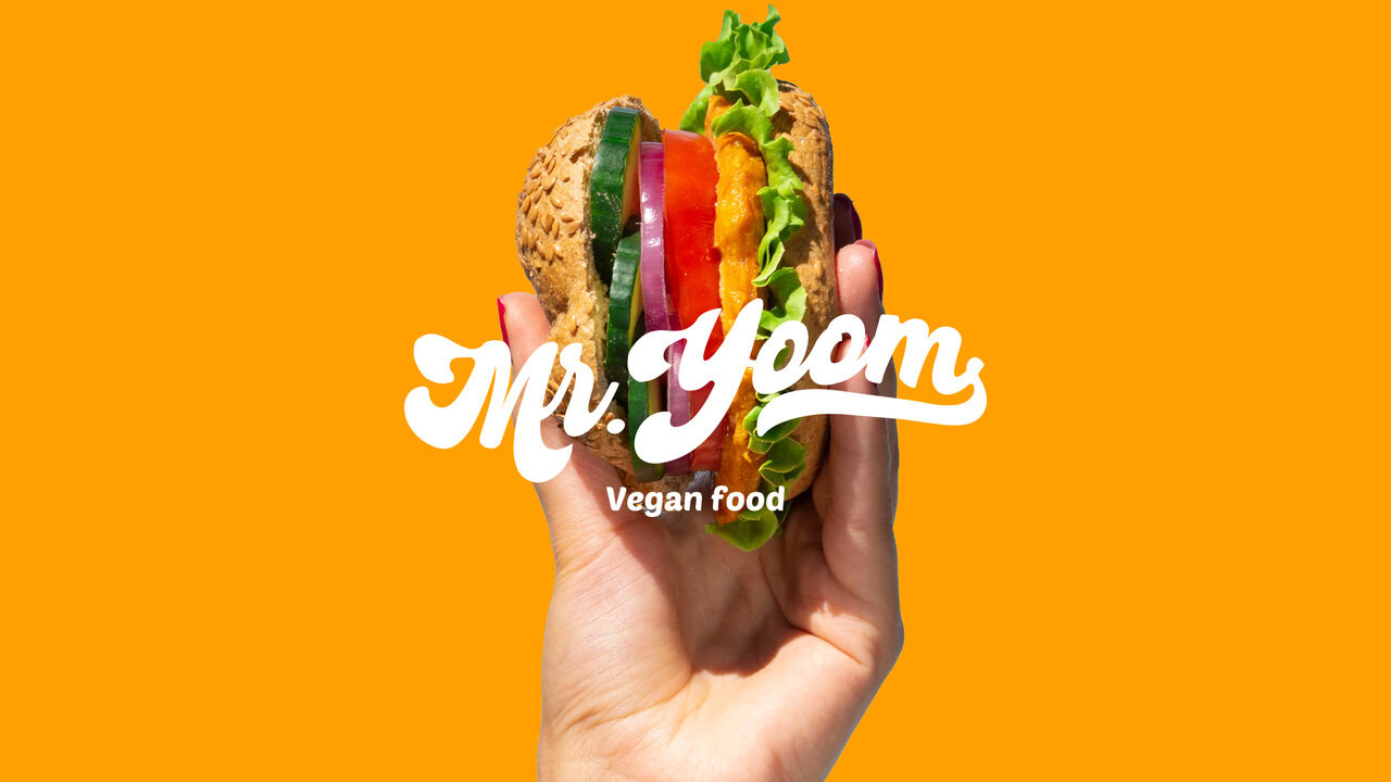 Nebly-Mr-Yoom-Logotipo Mano sosteniendo una hamburguesa vegana con el logotipo de Mr. Yoom y el texto 'Vegan food' superpuestos. Fondo de color naranja sólido. Diseño publicitario para comida rápida vegana.
