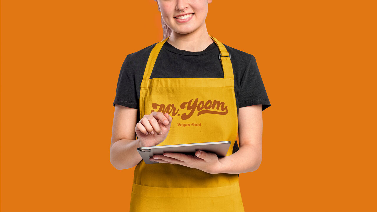 Nebly-Mr-Yoom-uniforme Mujer joven con un delantal amarillo que tiene el logo de mr,yoom en el frente, con un fondo color naranja.
