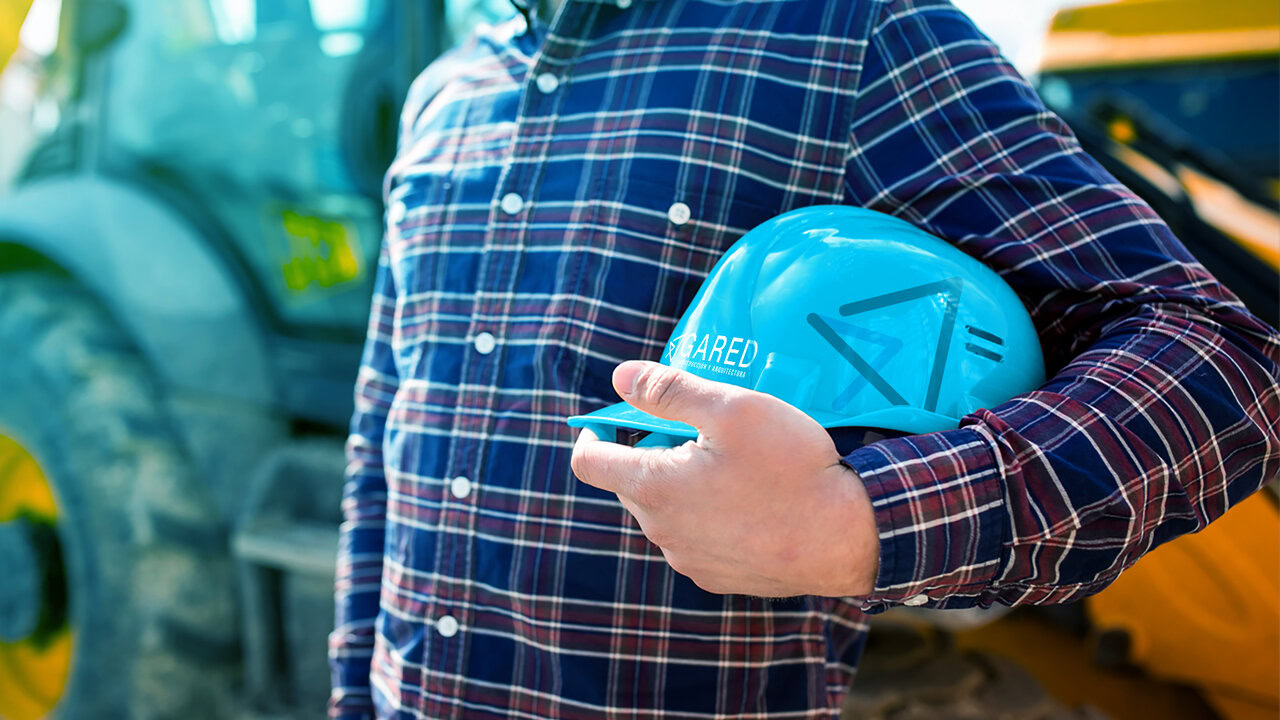 Nebly-logotipos-gared-portada Persona con camisa a cuadros sosteniendo un casco de construcción azul con el logotipo de Gared. Al fondo, maquinaria de construcción. Identidad minimalista para despacho de construcción: enfoque innovador y moderno.